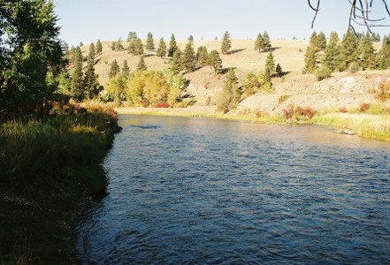 [The river is more like a wide stream at this location. There is a hillside across the river with some evergreens, but mostly dirt and stone.]